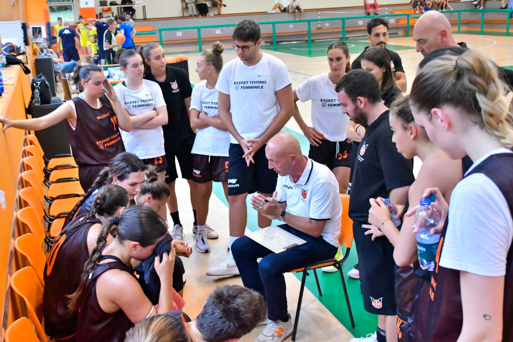 La squadra del Basket Torino Femminile riunita durante un timeout, con l'allenatore che fornisce indicazioni tattiche. Le giocatrici ascoltano attentamente mentre si preparano per il proseguimento della partita. Lo sfondo mostra il campo e l'ambiente di gioco.